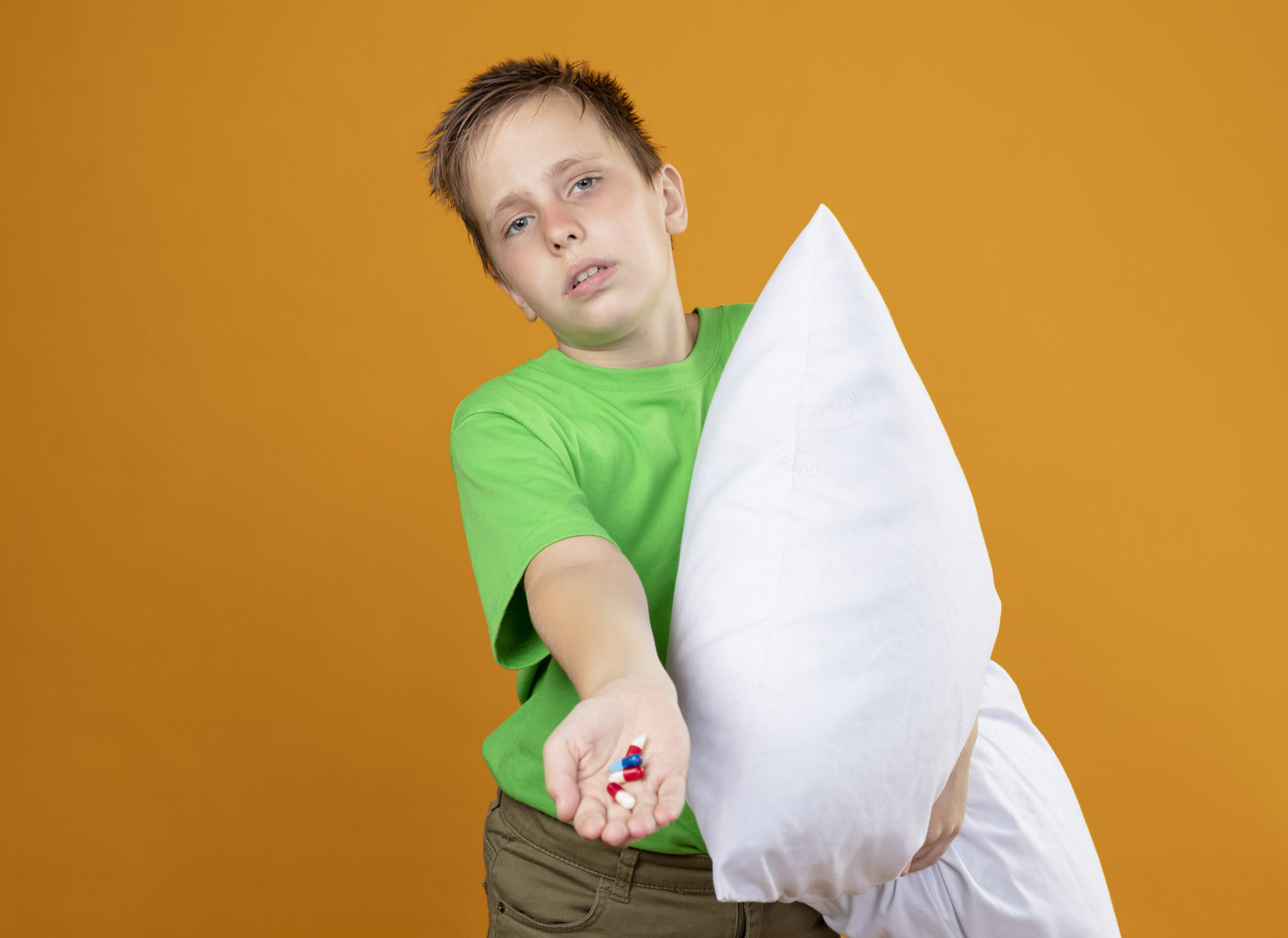 Ill little boy in green t-shirt feeling unwell holding pillow showing pills in hand looking confused unhappy and sick standing over orange wall